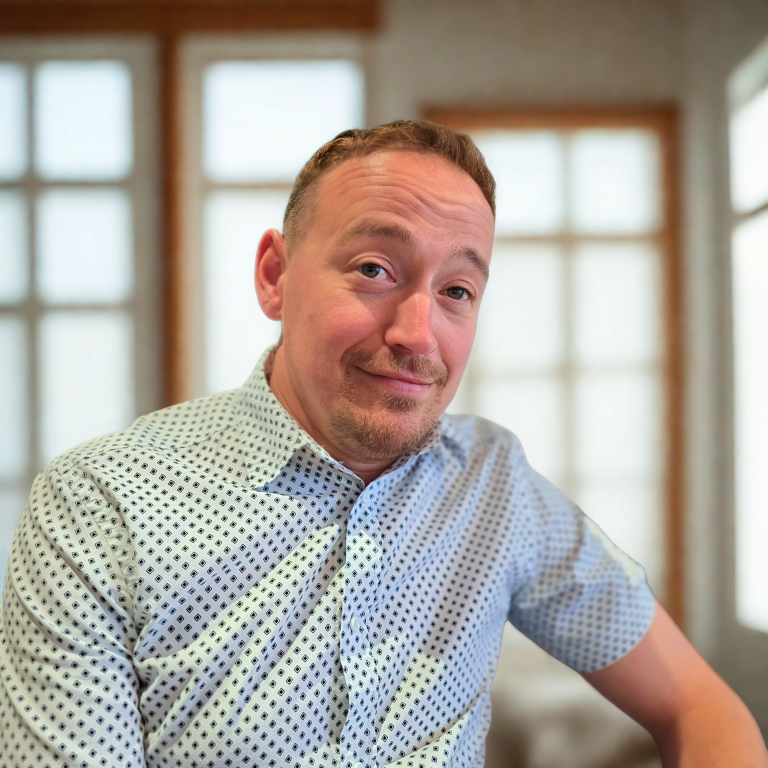 Homme souriant portant une chemise à motifs, assis dans un intérieur lumineux.